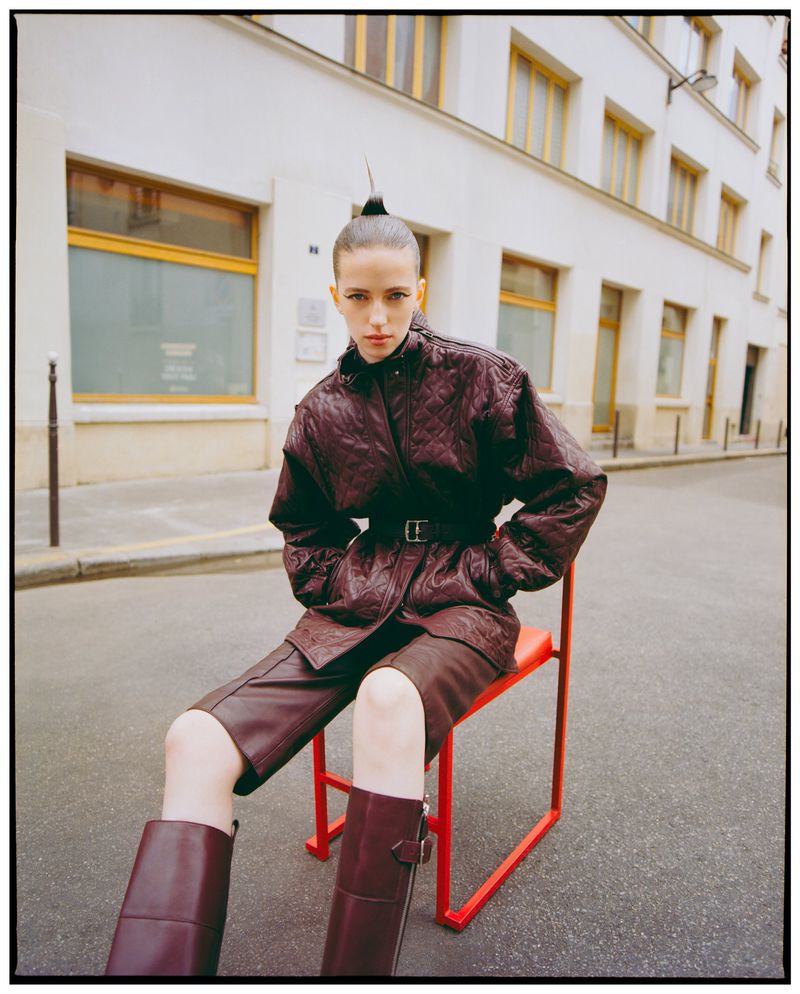 Fashion photo of a woman with unique updo sitting on a bright red chair on a street in a quilted jacket and boots.