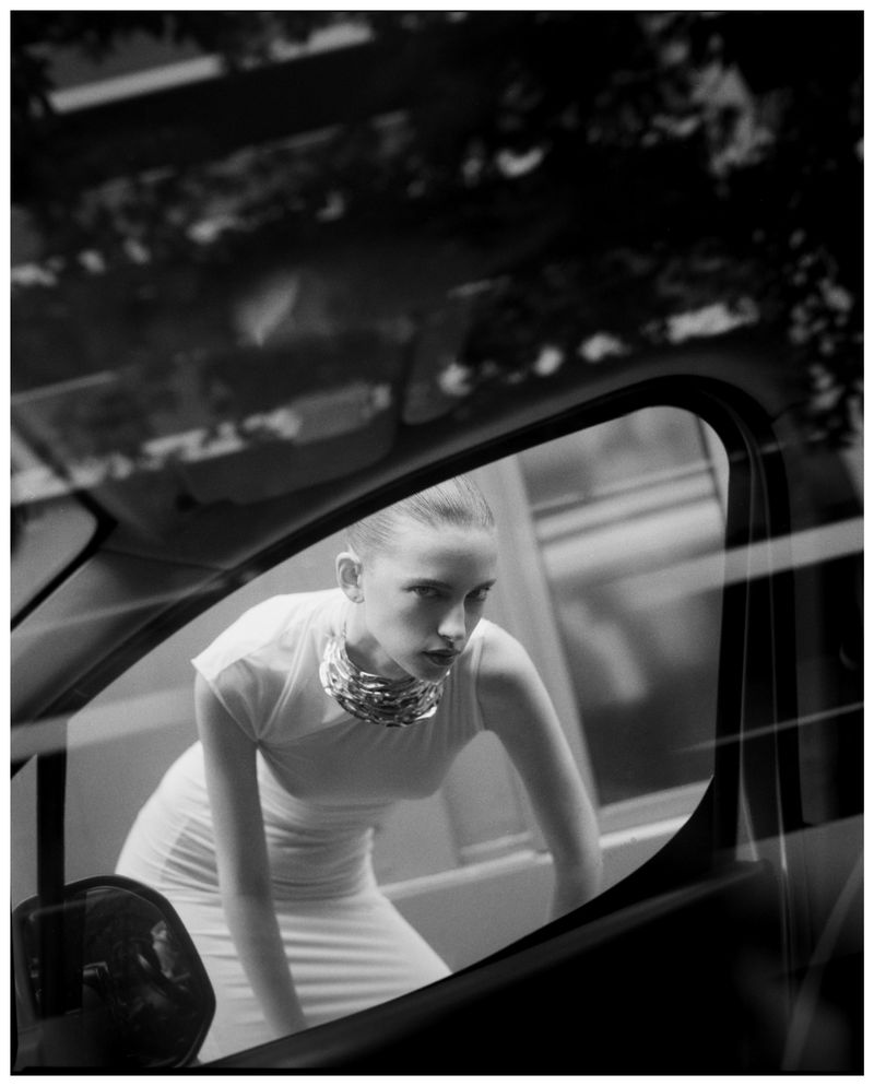 Black-and-white fashion shot of a slender woman in a white dress and chunky chain necklace, seen through the open window of a car.