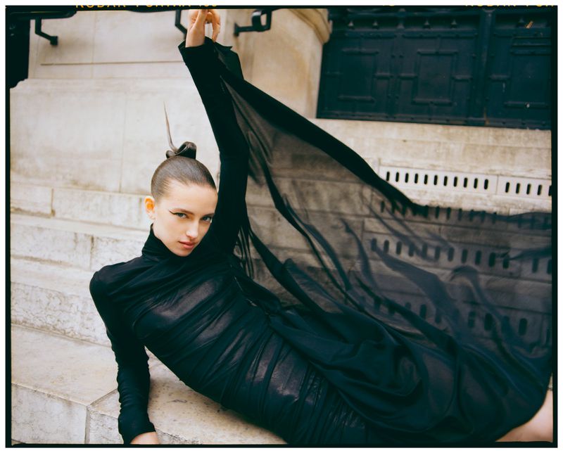 Woman in black dress and dramatic eye makeup sits on stone steps, one arm raised, sheer fabric flowing in urban outdoor fashion shot.