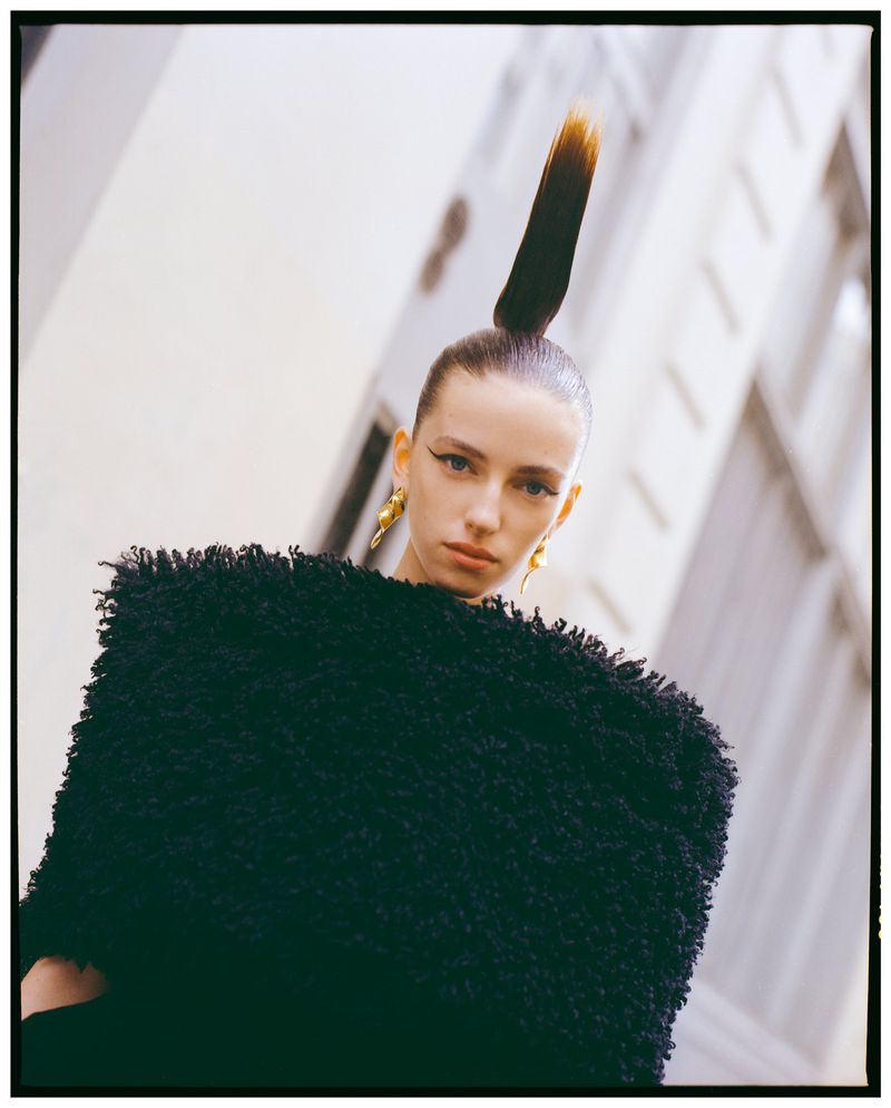 Portrait of a fair-skinned adult woman with a high ponytail, wearing a black textured top, against a white building backdrop.