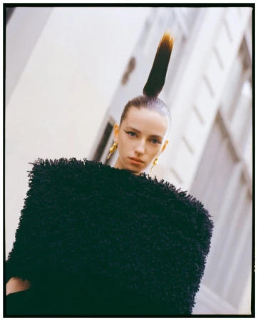 Portrait of a fair-skinned adult woman with a high ponytail, wearing a black textured top, against a white building backdrop.