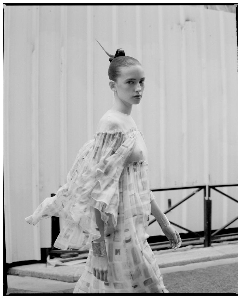 Monochrome portrait of a poised, fair-skinned adult woman in a billowing, patterned dress, set against a stark white wall, editorial fashion.