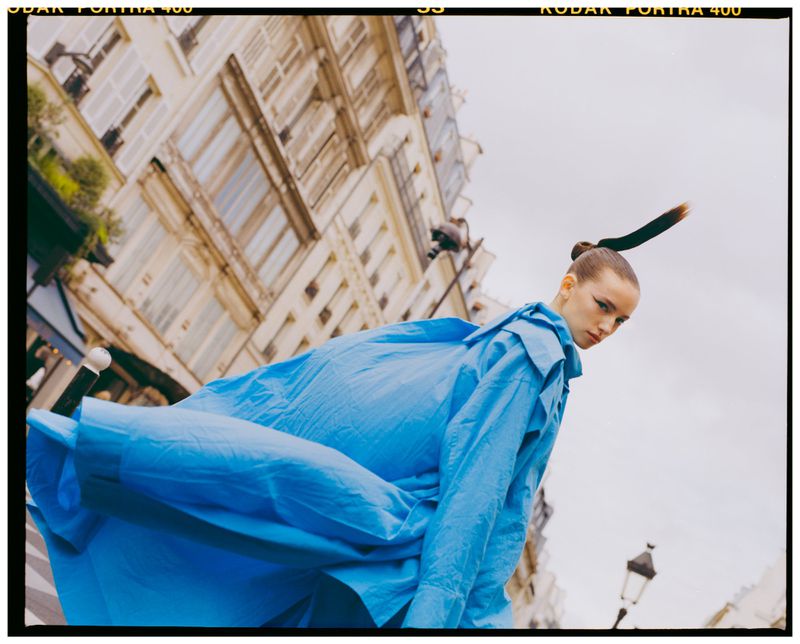 Model in vibrant blue coat captured in a low-angle street view with traditional European architecture against an overcast sky.