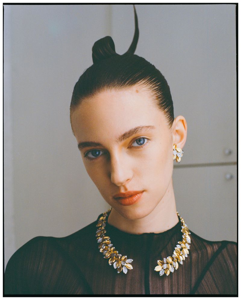Close-up portrait of a woman with a unique updo, wearing a leaf-patterned necklace and earrings against a light grey background.