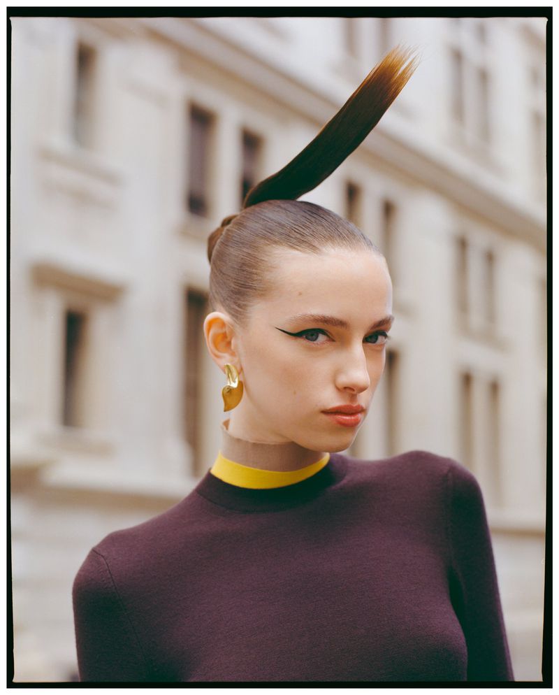 Close-up portrait of a woman with sleek hair and a feather extension, wearing a dark top with a yellow collar, outdoors.