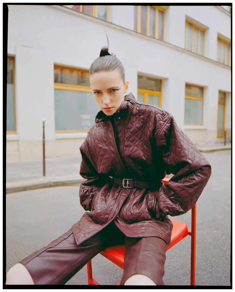 Fashion portrait of an adult female wearing a burgundy quilted jacket and pants sitting on a red chair in an urban environment.