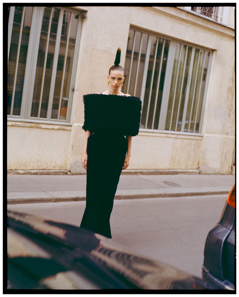 Model standing on a city street wearing a structured black top with sculptural shoulders and high ponytail hairstyle