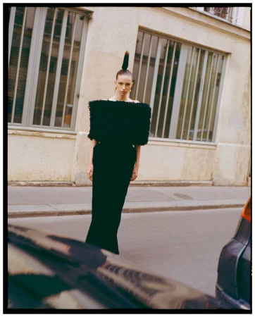 Model standing on a city street wearing a structured black top with sculptural shoulders and high ponytail hairstyle