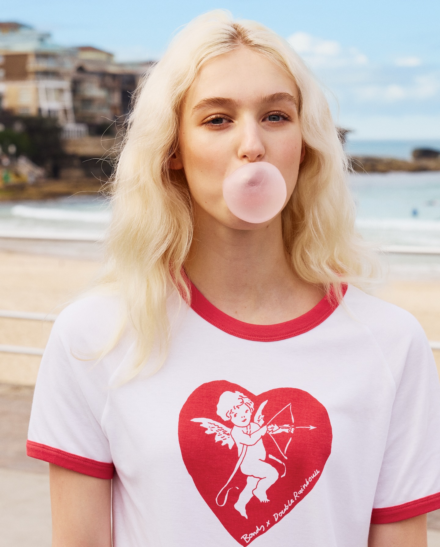A young woman with platinum blonde hair blows a pink bubblegum bubble while standing on a beach with buildings in the background.
