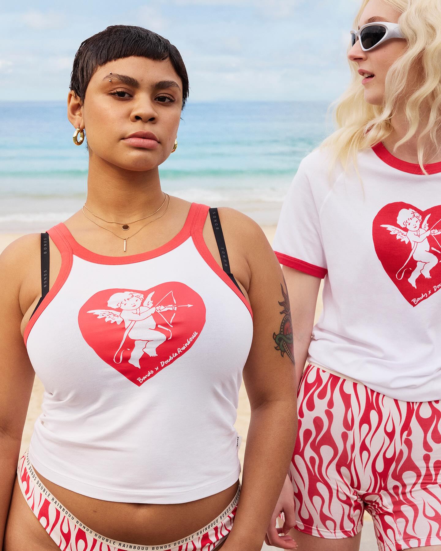 Two women stand on a beach wearing white tops with cupid heart designs and patterned bottoms, showcasing a bright, summery fashion style.