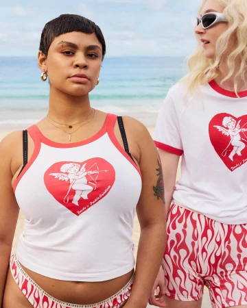 Two women stand on a beach wearing white tops with cupid heart designs and patterned bottoms, showcasing a bright, summery fashion style.
