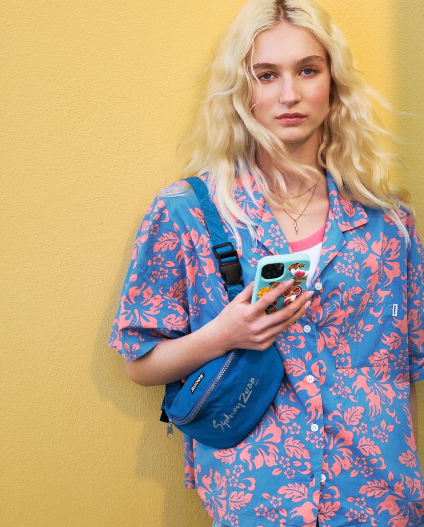 Woman with wavy blonde hair poses in a blue floral shirt with a matching blue waist bag against a yellow background, holding a phone.