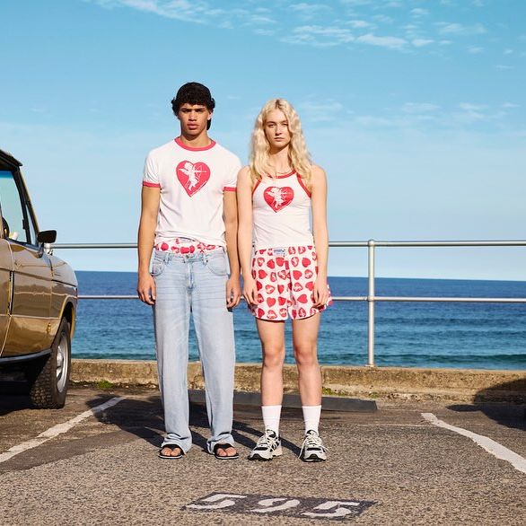 Two people, male and female, stand in an outdoor parking lot near the ocean, wearing matching heart-themed outfits by a gold SUV.