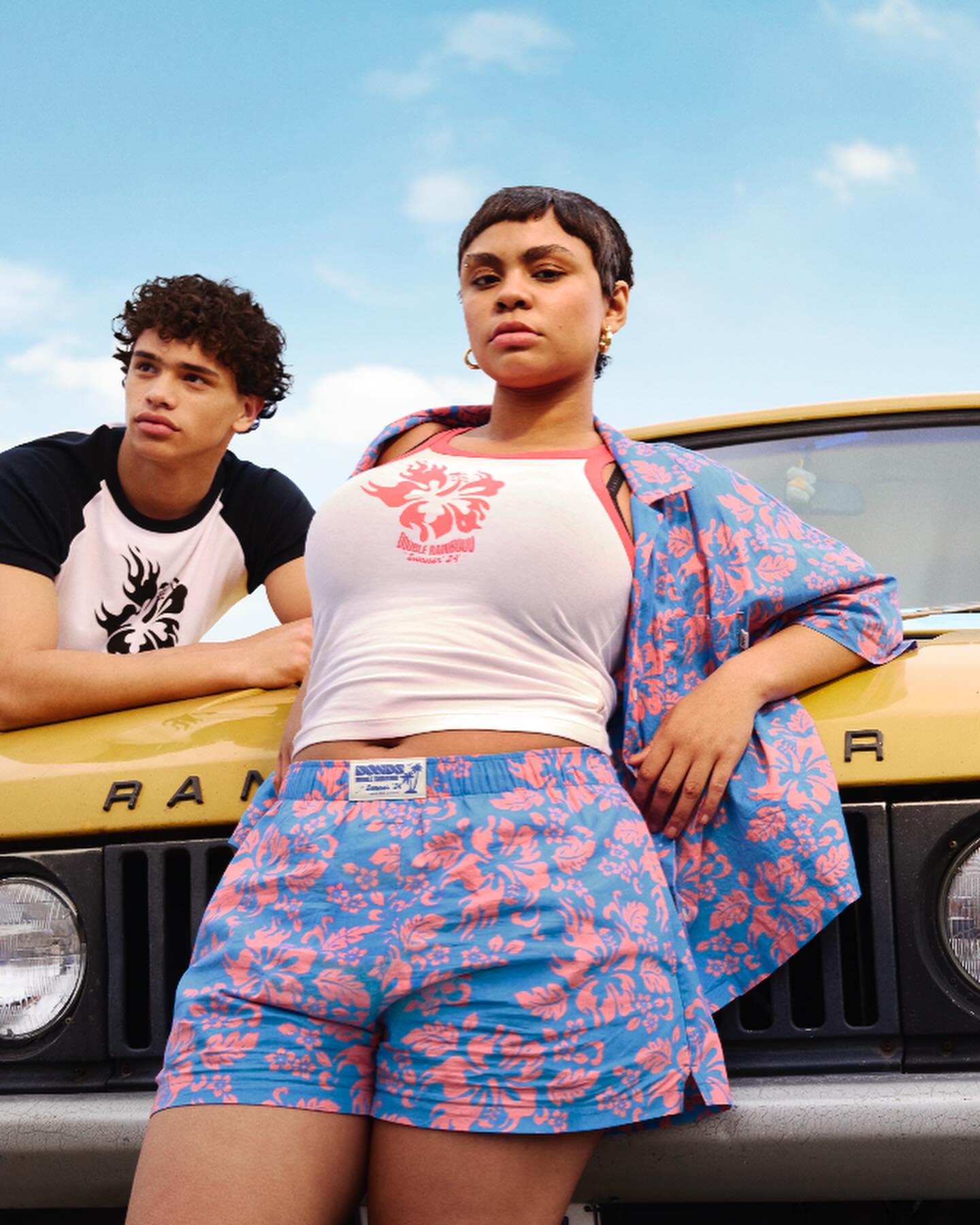 Two fashion models pose near a yellow vehicle wearing matching blue and pink tropical print shorts and shirts in an outdoor setting.