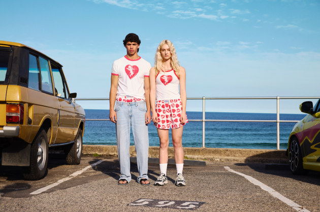 Two fashion models stand in front of vintage cars at a beach parking lot wearing matching white shirts with red heart graphics.