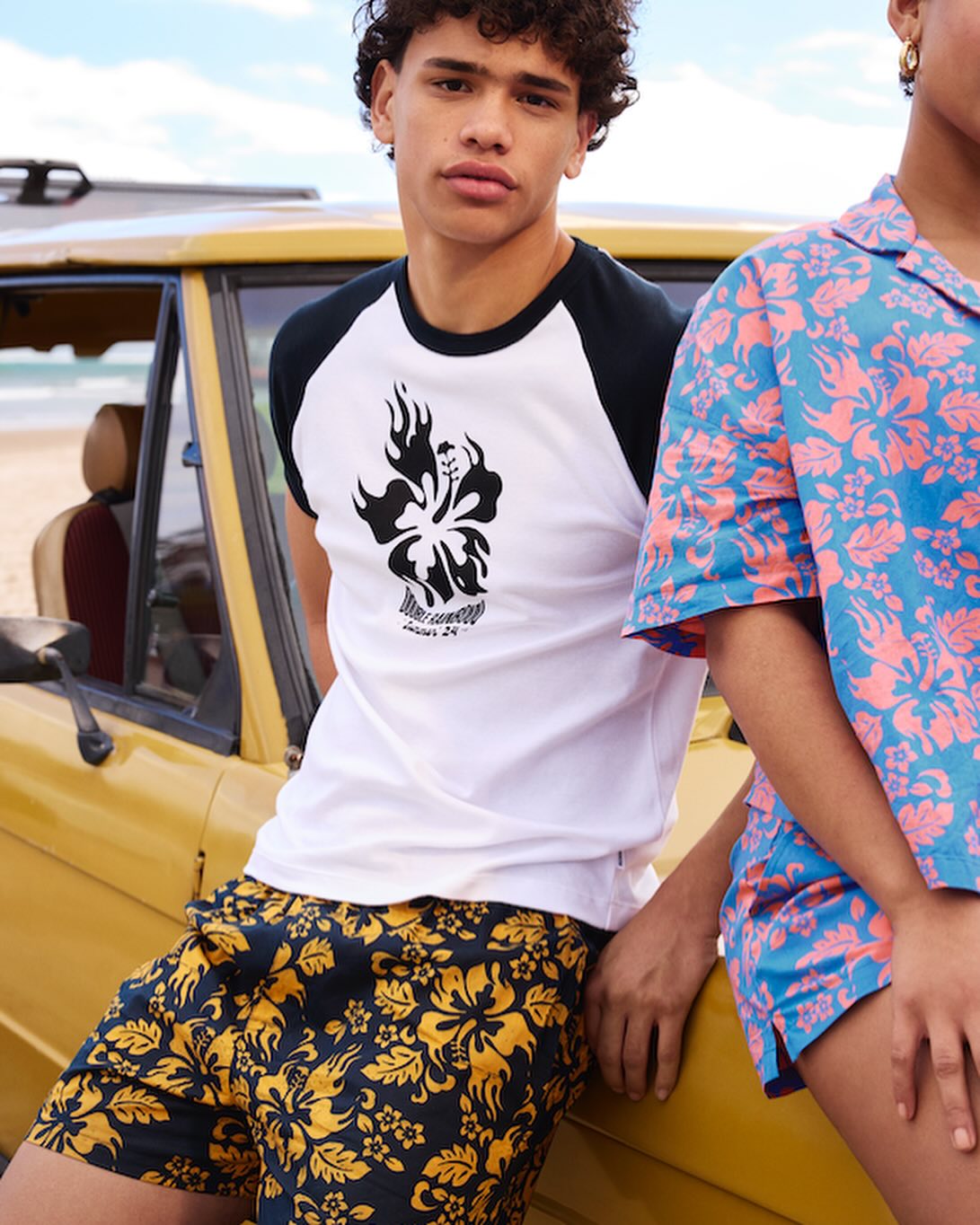 Two people in patterned shirts and shorts pose next to a vintage car on a beach. One person wears a t-shirt with a floral skull.