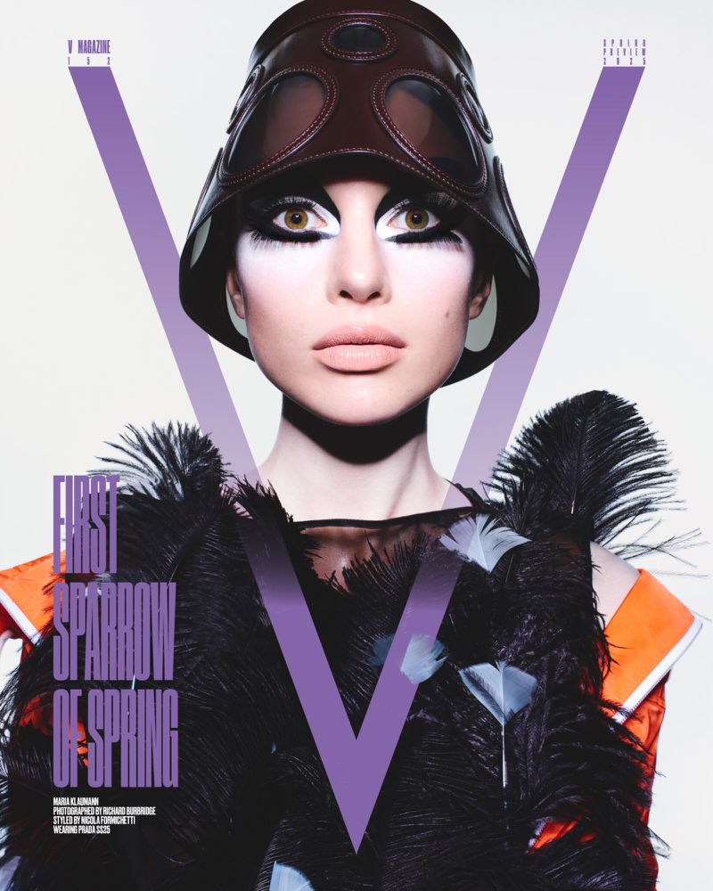 Close-up shot of a fair-skinned woman wearing a leather helmet and dramatic eye makeup on a V Magazine cover. Feathers adorn the garment.