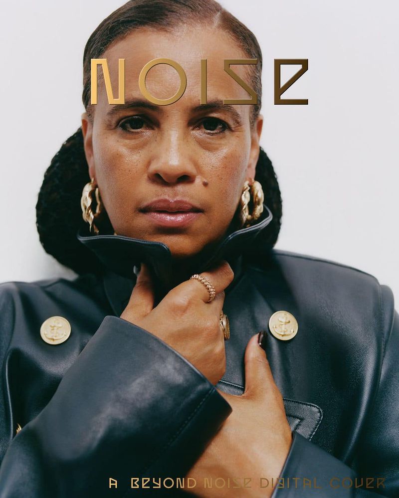 Portrait of a woman with gold earrings and a black jacket, overlaid with the gold text "NOISE," against a plain background.