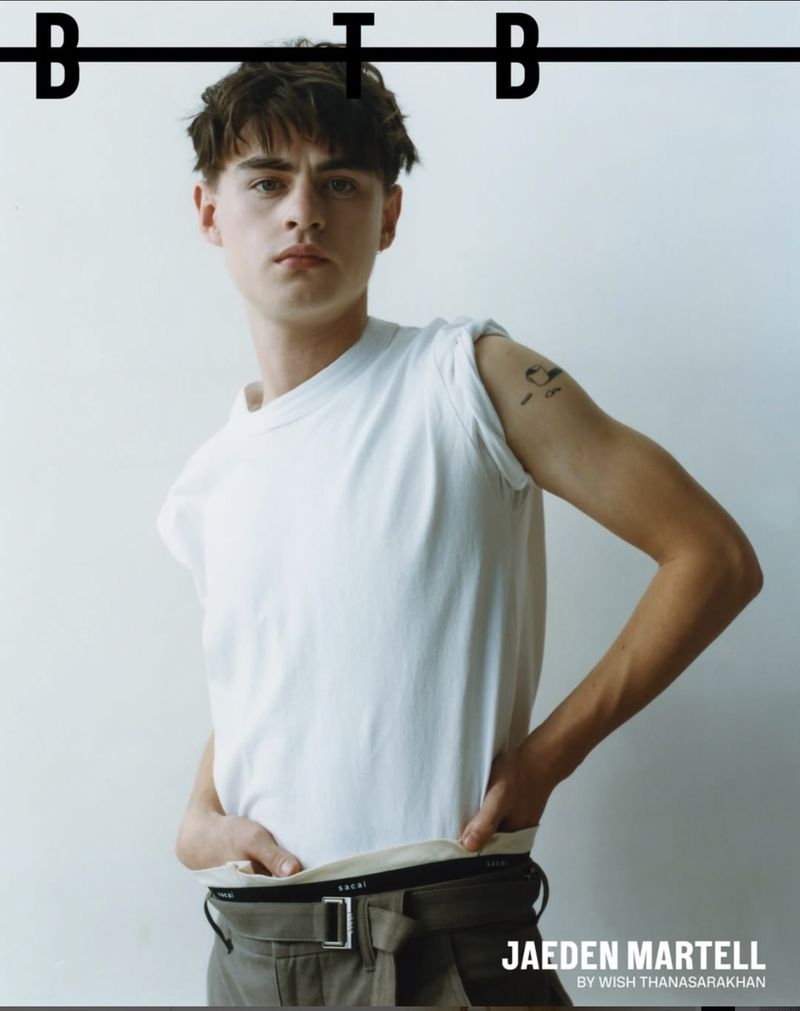 A young man in a white T-shirt poses indoors against a white wall, wearing dark pants with a belt, captured in a medium shot.