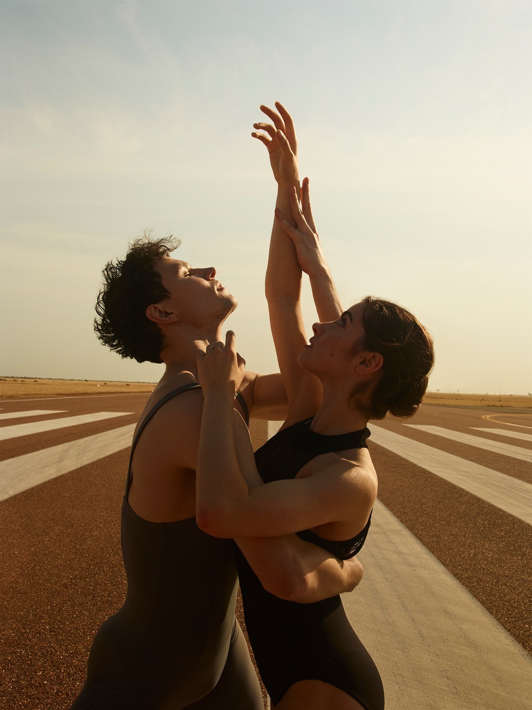 Two ballet dancers, an adult male and female, perform together outdoors on a runway with the sky in the background in a medium shot.