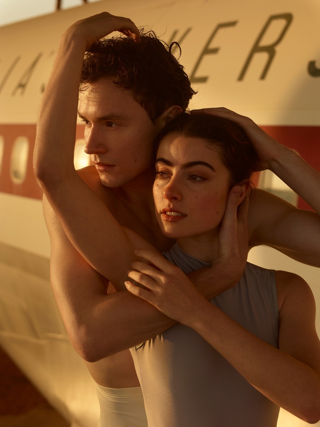 Close-up of a couple embraced in front of an airplane, soft focus. The male figure is bare-chested and the female wears a leotard.