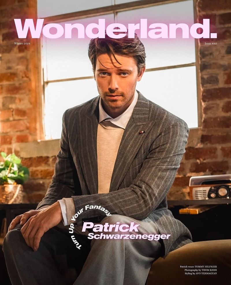 Patrick Schwarzenegger poses for a magazine cover, wearing a gray striped suit and a neutral sweater, seated indoors near a window.