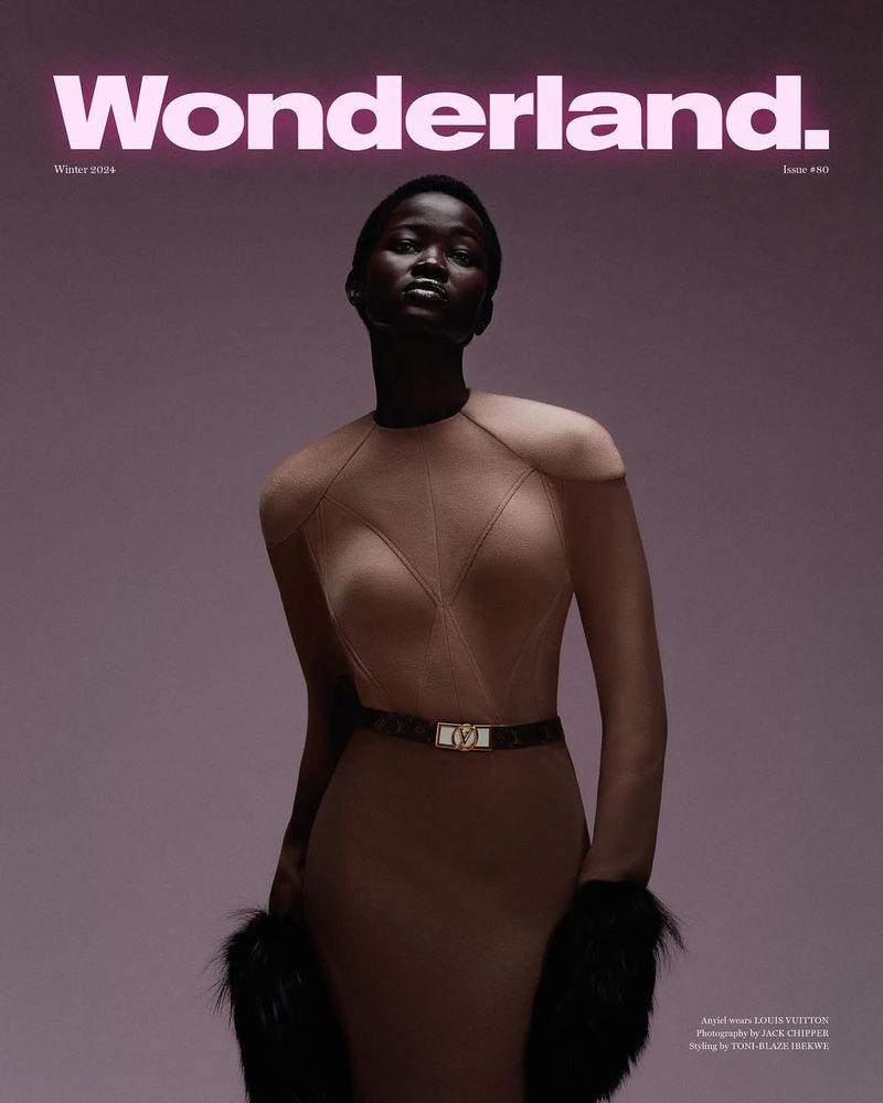 Magazine cover featuring a dark-skinned woman in a beige dress with fur wrist trim and brown belt against a mauve background.