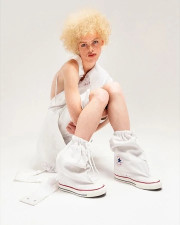Person with blond afro hair wearing a white dress and white sneaker boots sitting in a photo studio against a white background.