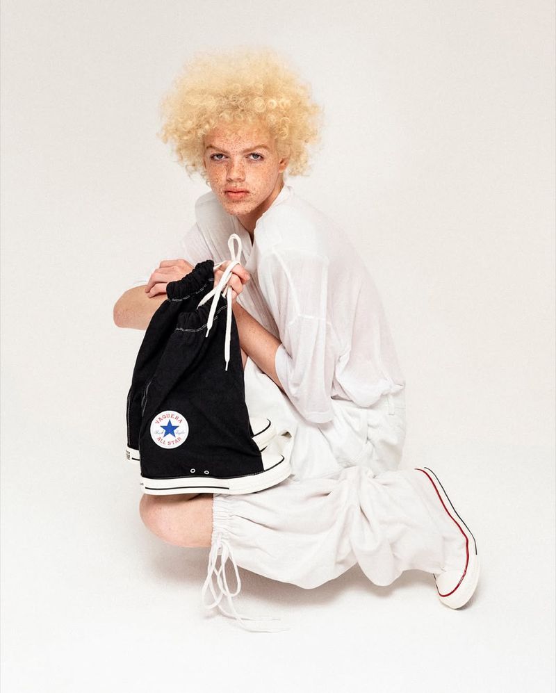 A person with an afro squats on white background holding a Converse shoe bag, wearing white shirt and ruched pants, fashion shot.
