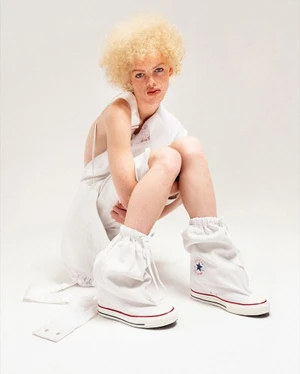 Model with blonde curly hair crouching in a white top and oversized white fabric Converse sneakers against a seamless white studio background.