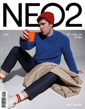 Man wearing a blue sweater and red beanie holding an orange cup sitting on a grey floor for a magazine cover
