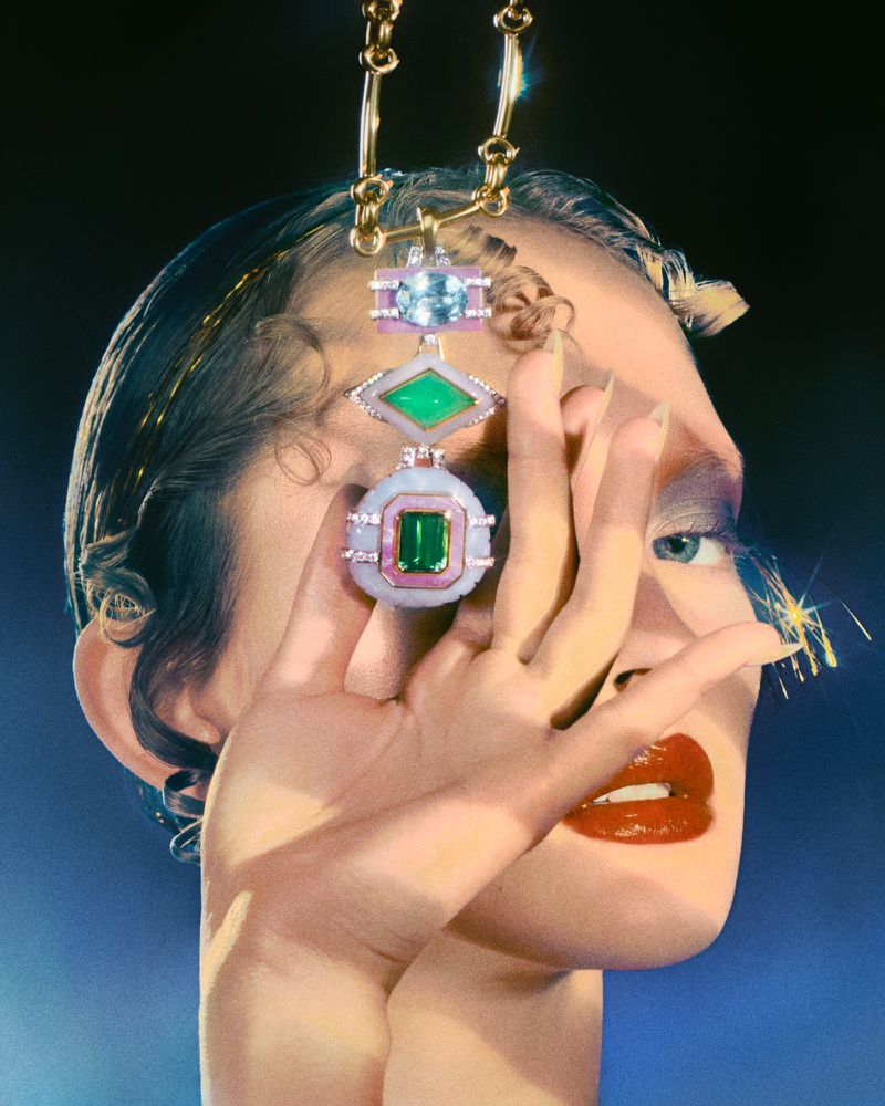 Fashion photo featuring a woman's face partially obscured by her hand, adorned with red lipstick, blue eyeshadow, and a large gold pendant necklace.