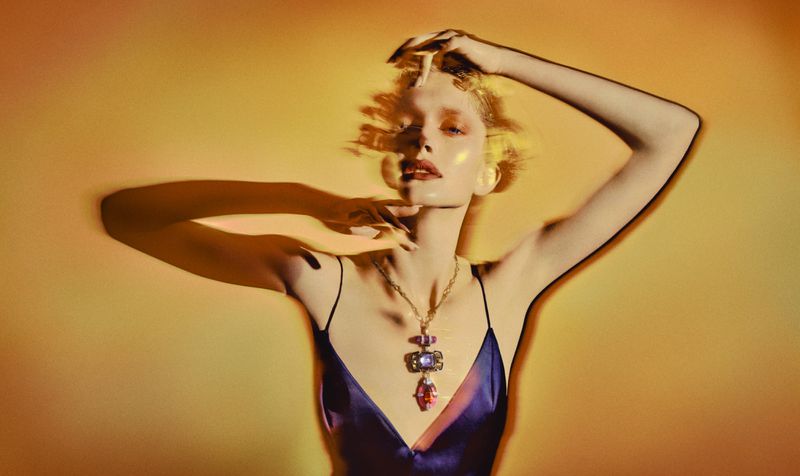 Woman with hands raised in a photo studio with warm lighting, wearing a purple slip dress and jewelry posing against a yellow backdrop.