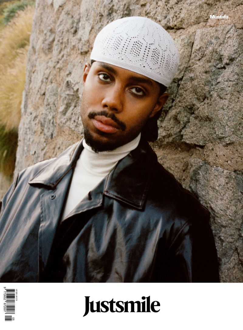 Portrait of a man with a white cap and dark coat leaning against a stone wall, with visible text reading 'Justsmile' on a magazine cover.