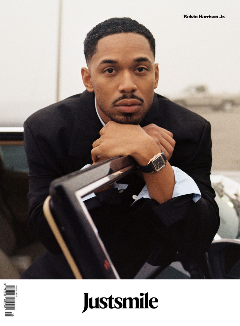 Portrait of a young African-American man in a black suit and watch, posing with hands clasped on the top of a black car.