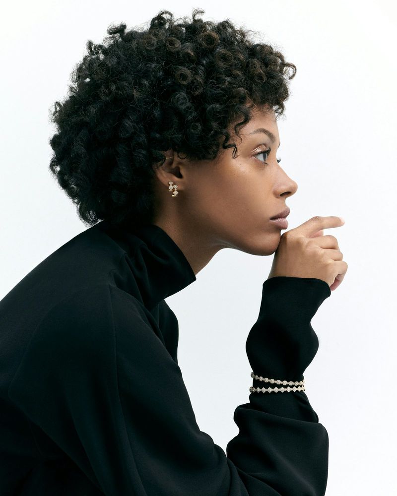 Profile view of a woman with curly hair and neutral expression, wearing a black turtleneck with layered pearl bracelets against white backdrop.