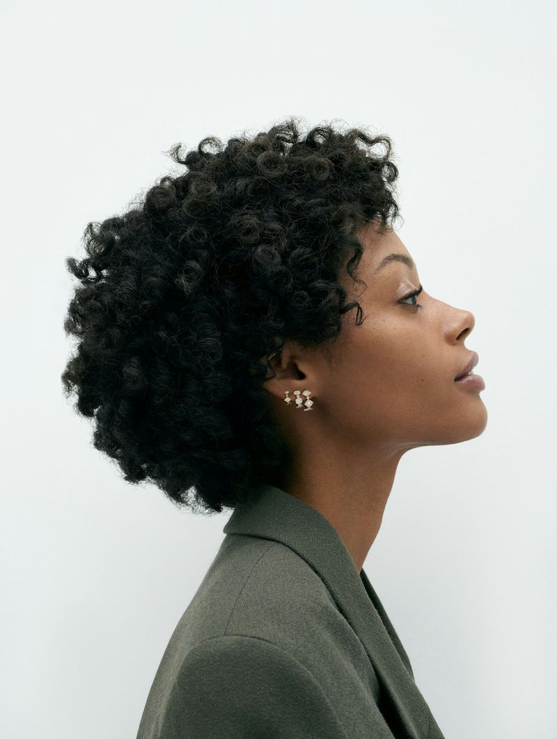 Profile portrait of a woman with curly hair wearing a gray-green jacket and gold earrings against a bright white background.