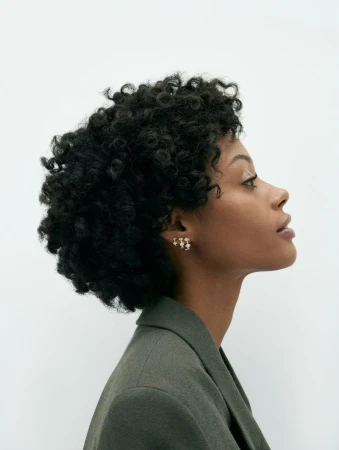 Profile portrait of a woman with curly hair wearing a gray-green jacket and gold earrings against a bright white background.