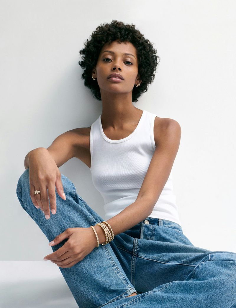 A woman with curly hair sits against a white wall, wearing a white tank top and blue jeans, accessorized with bracelets and a ring.