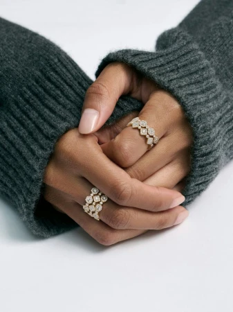 Close-up of hands in a dark grey knit sweater wearing gold and diamond rings from David Morris against a white surface.