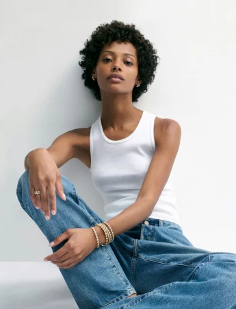 Model Londone Myers seated in a white tank top and blue jeans wearing gold jewelry against a white studio backdrop.