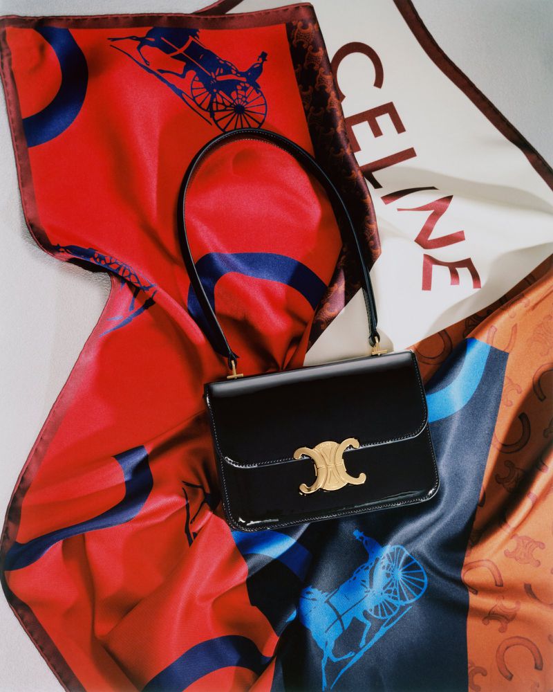 A black leather Celine handbag with a gold logo, lying on a colorful patterned silk scarf. Studio lit product shot.