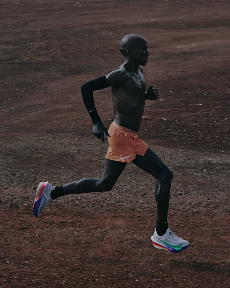 Side profile shot of a dark-skinned adult male runner in motion wearing orange shorts and blue and white running shoes on a dirt track.