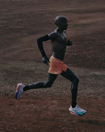 Side profile shot of a dark-skinned adult male runner in motion wearing orange shorts and blue and white running shoes on a dirt track.