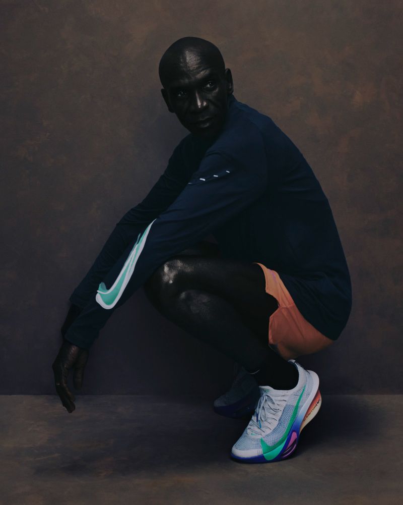 Athletic Black man in a squat wearing a long-sleeved blue Nike shirt and colorful running shoes against a textured brown backdrop.