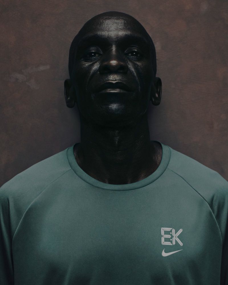 Close-up portrait of a Black adult male wearing a teal t-shirt with a white logo, set against a textured gray background.