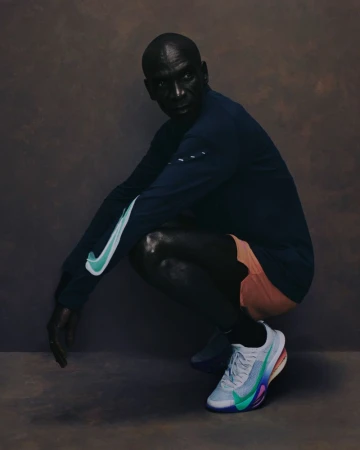 Eliud Kipchoge squatting in a dark long-sleeve shirt, orange shorts, and running shoes against a textured studio backdrop