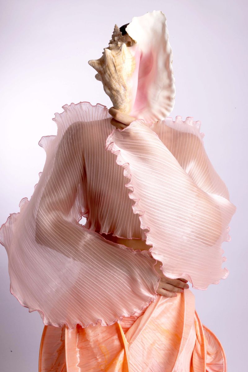 Person in a translucent ruffled blouse holding a large conch shell over their face against a plain backdrop, creating a surreal effect.