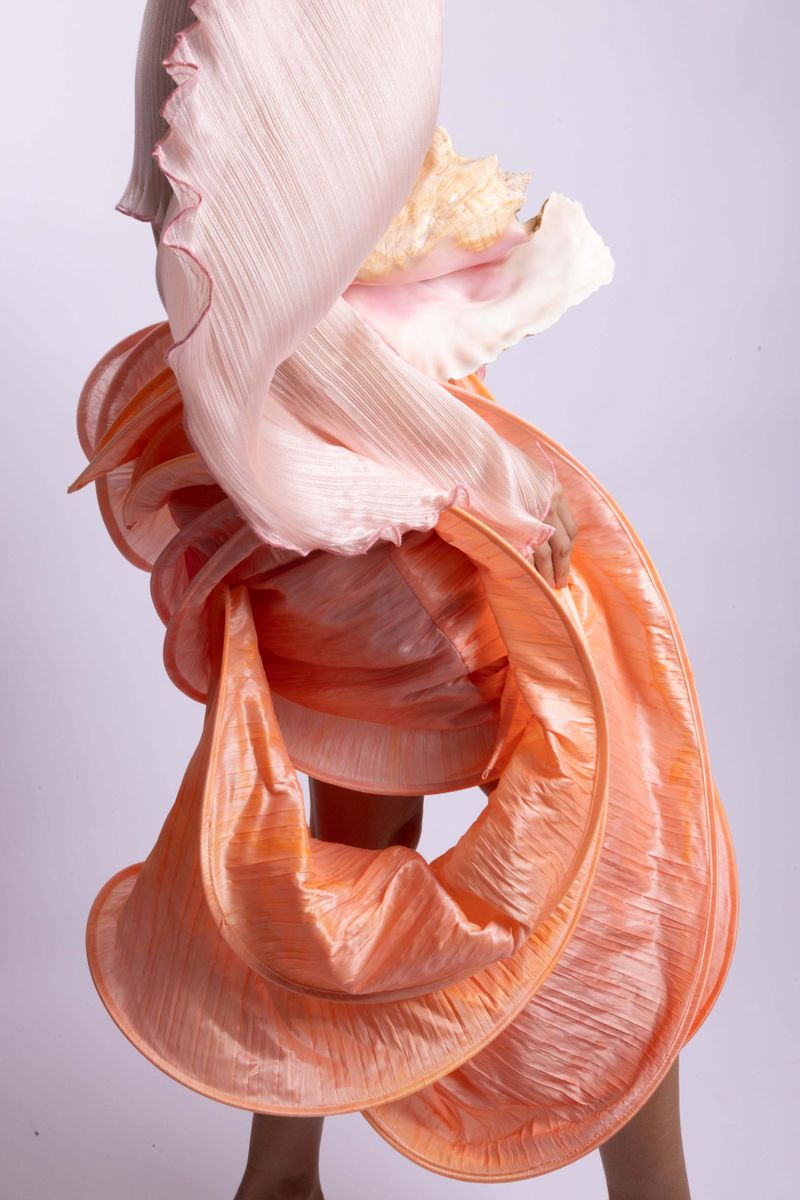 Model wearing a pleated sculptural spiral peach garment and pale pink draping against a plain light studio backdrop.