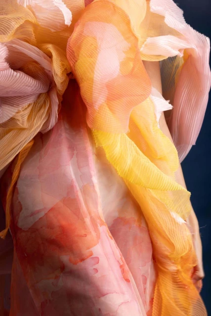 Close-up of flowing pleated fabrics in shades of pink, yellow, and orange against a dark blue background, capturing a soft and ethereal aesthetic.